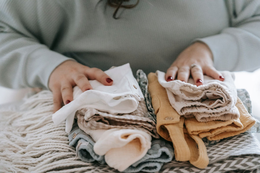 Hands arranging folded baby clothes in neutral colours | يدان ترتّبان ملابس بيبي مطوية بألوان هادئة