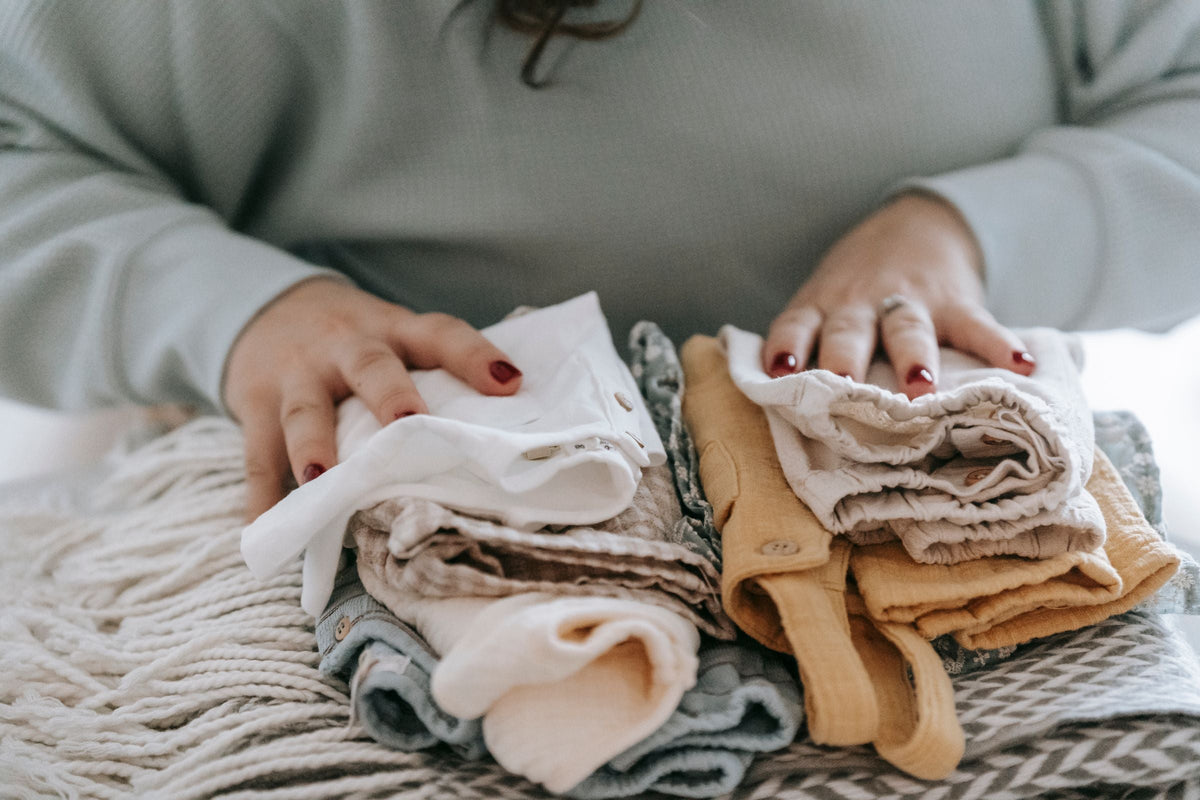 Hands arranging folded baby clothes in neutral colours | يدان ترتّبان ملابس بيبي مطوية بألوان هادئة